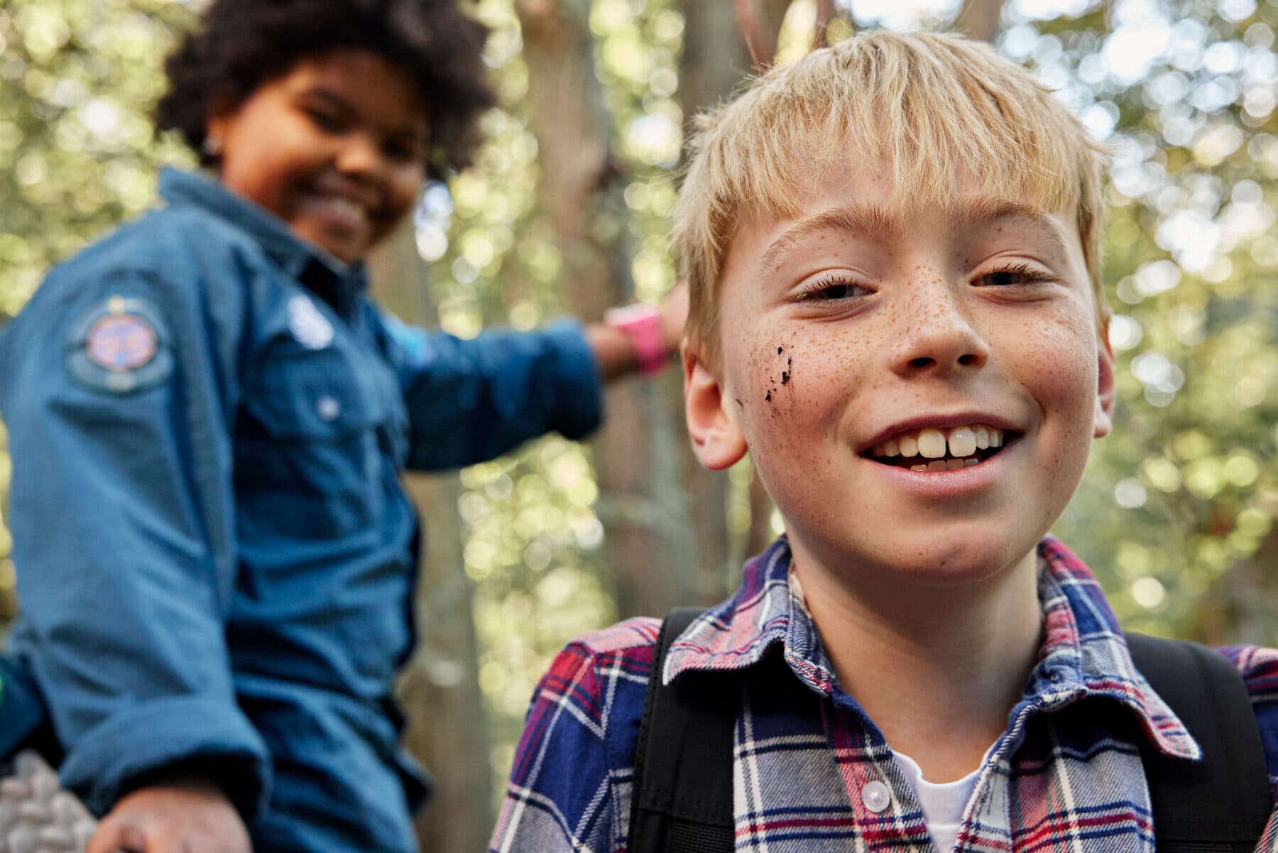 Två scouter tillsammans ute i skogen. En scout syns i bakgrunden och har på sig scoutskjorta. En syns i förgrunden och har en rutig skjorta.