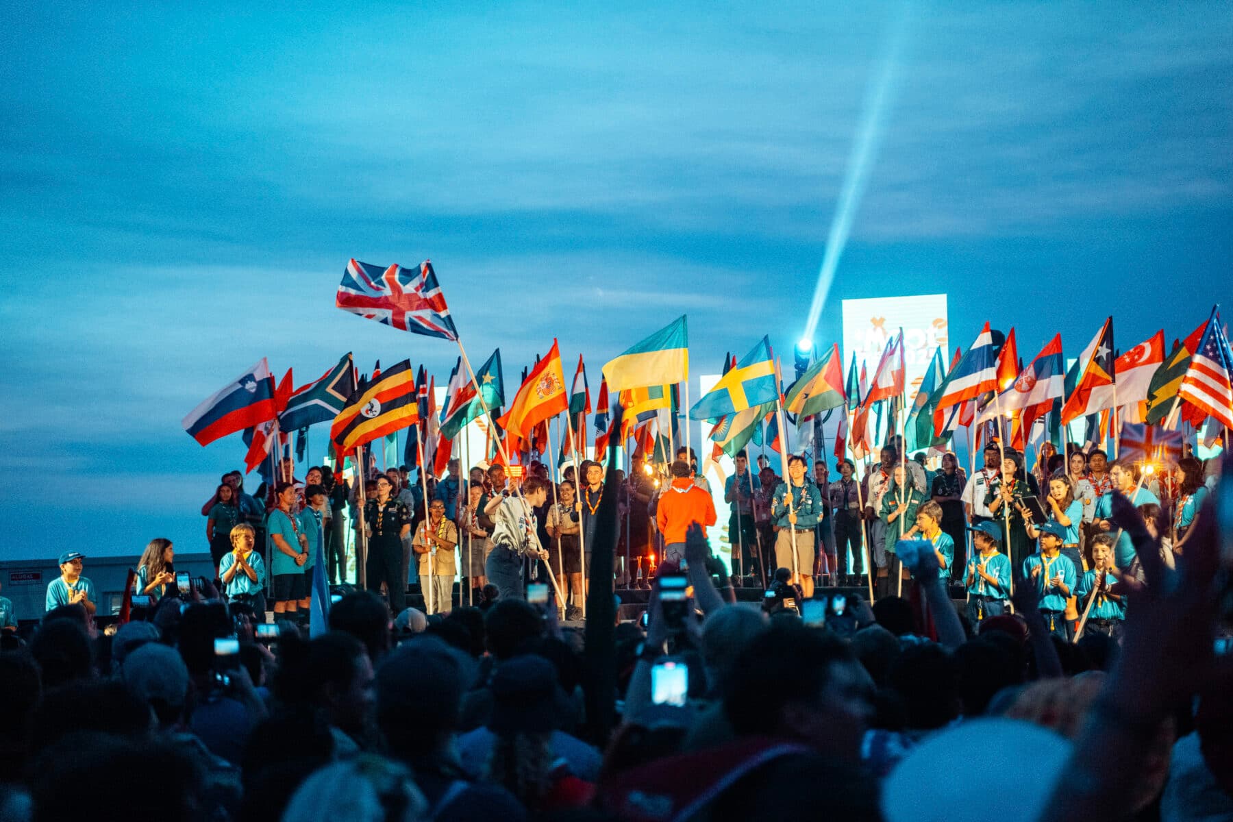 En grupp människor står på en scen, många håller internationella flaggor från olika länder. På scenen syns scoutuniformer, en brinnande fackla och en bakgrund med blå himmel och strålkastarljus.