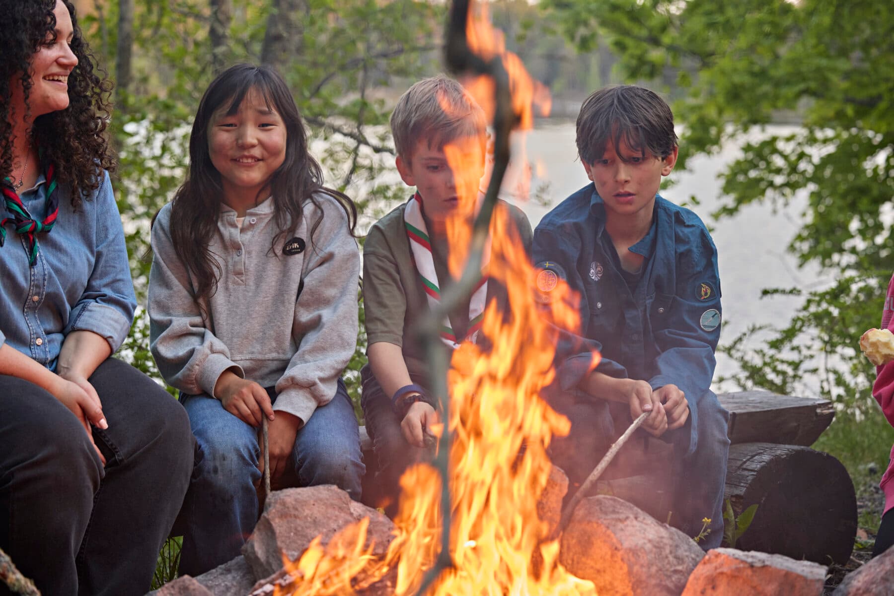 Personer sitter runt en lägereld i en skogsmiljö nära en sjö, med lågorna i förgrunden och omgivande träd i bakgrunden.