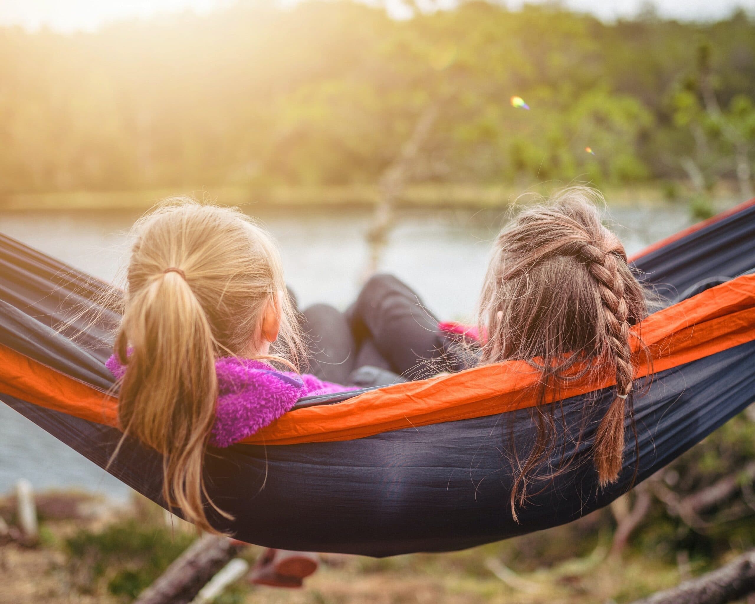 Två barn ligger i en orange och blå hängmatta nära en sjö, med skog i bakgrunden och varmt solljus som lyser igenom.