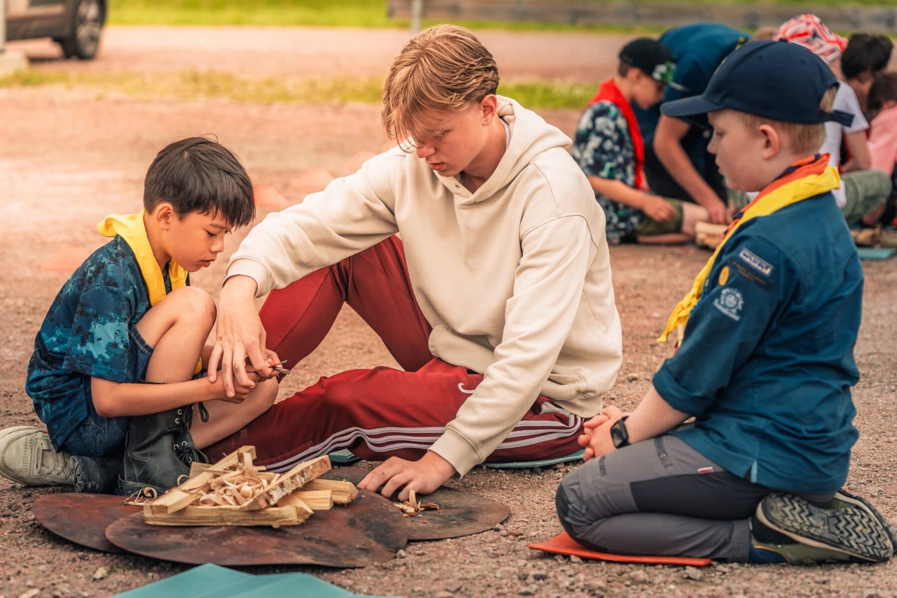 En grupp scouter sitter på marken och täljer träbitar tillsammans, med träspån samlade framför dem.
