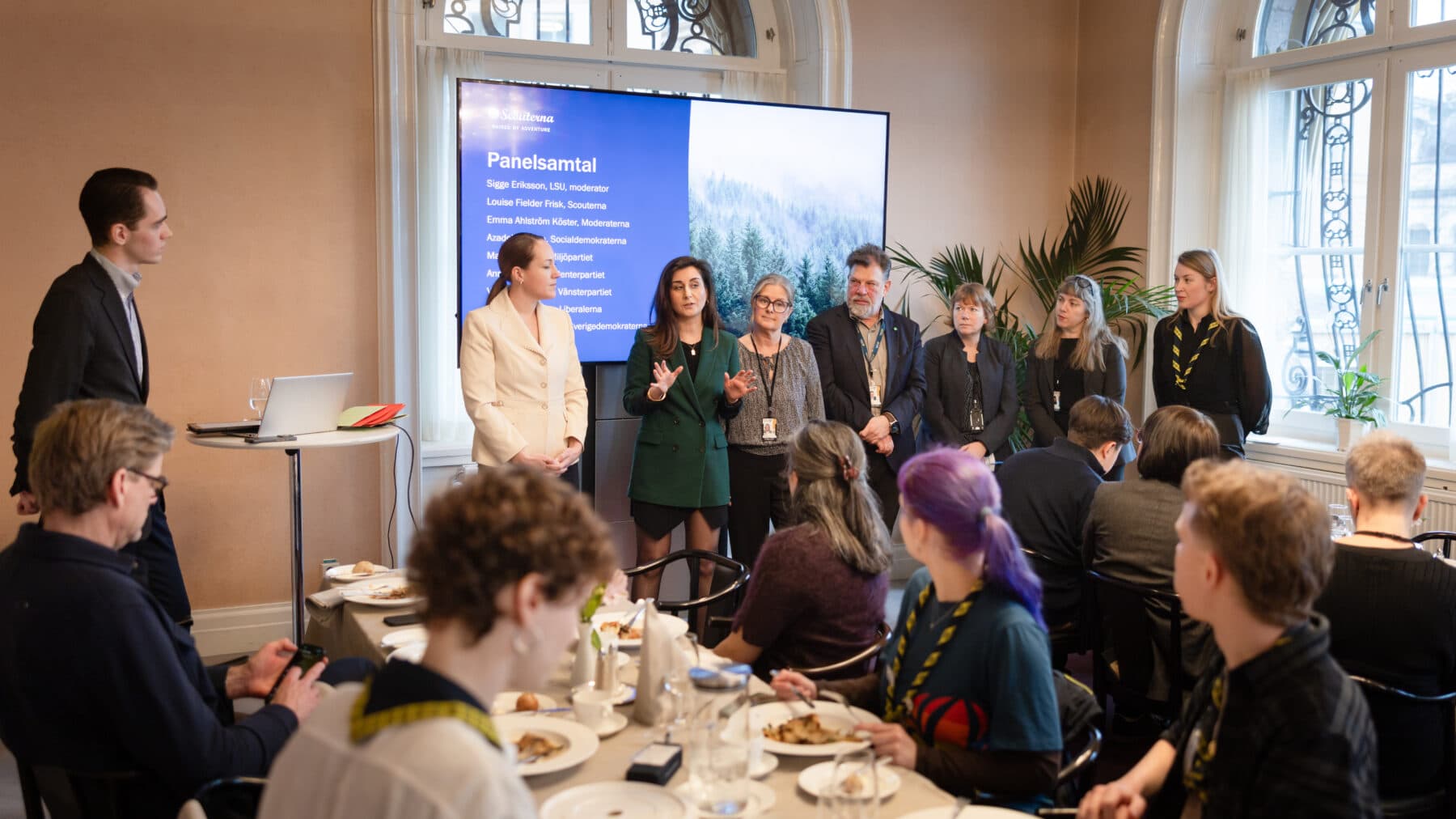 En paneldiskussion med flera personer som står framför en stor skärm med texten "Panelsamtal". Texten listar namn och roller, inklusive moderator och olika partiföreträdare. Publiken sitter vid bord med tallrikar och glas i förgrunden.