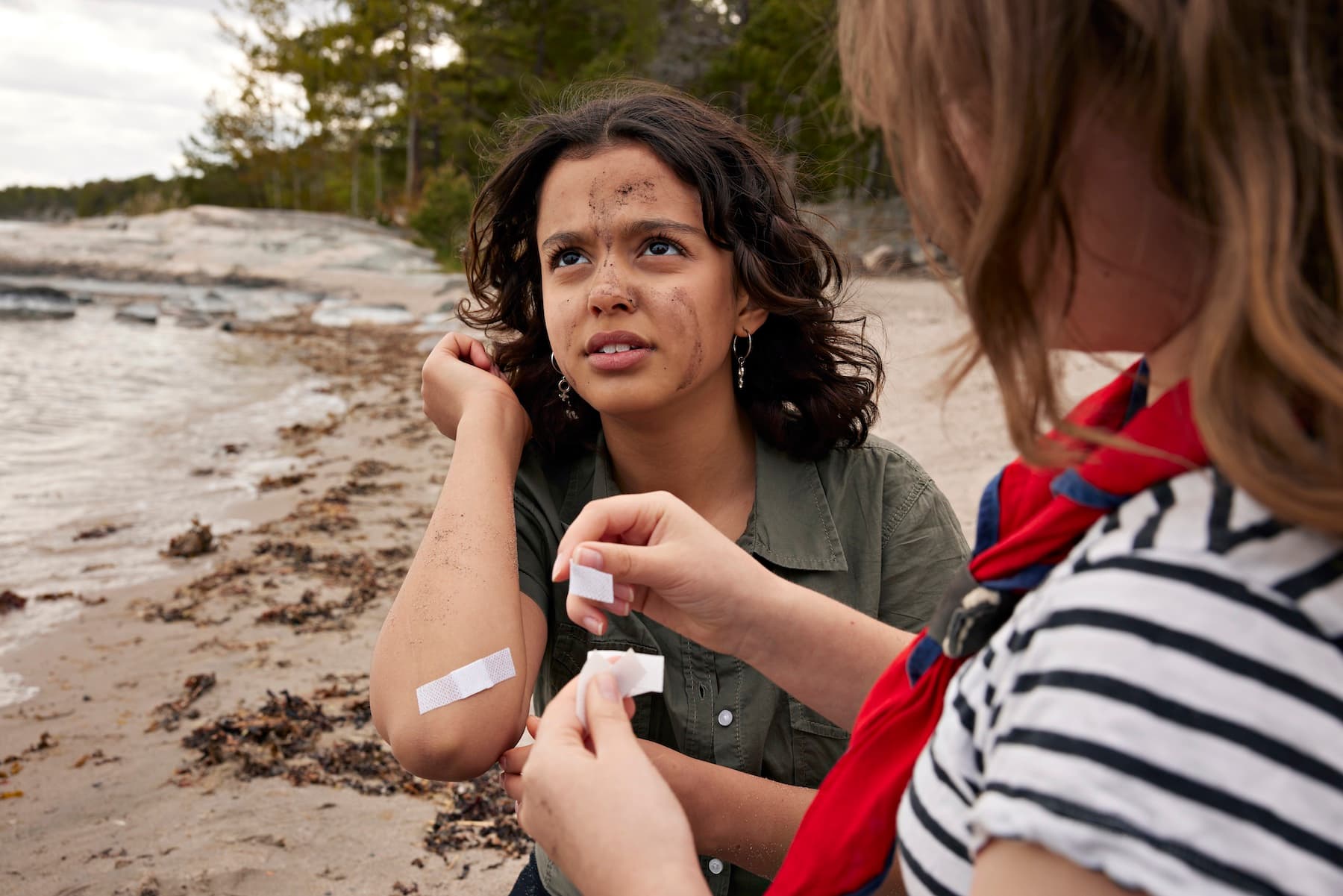 En person med mörkt lockigt hår sitter vid en strand och får hjälp av en annan person med att sätta plåster på en skadad arm. Bakgrunden visar sand, stenar och träd.