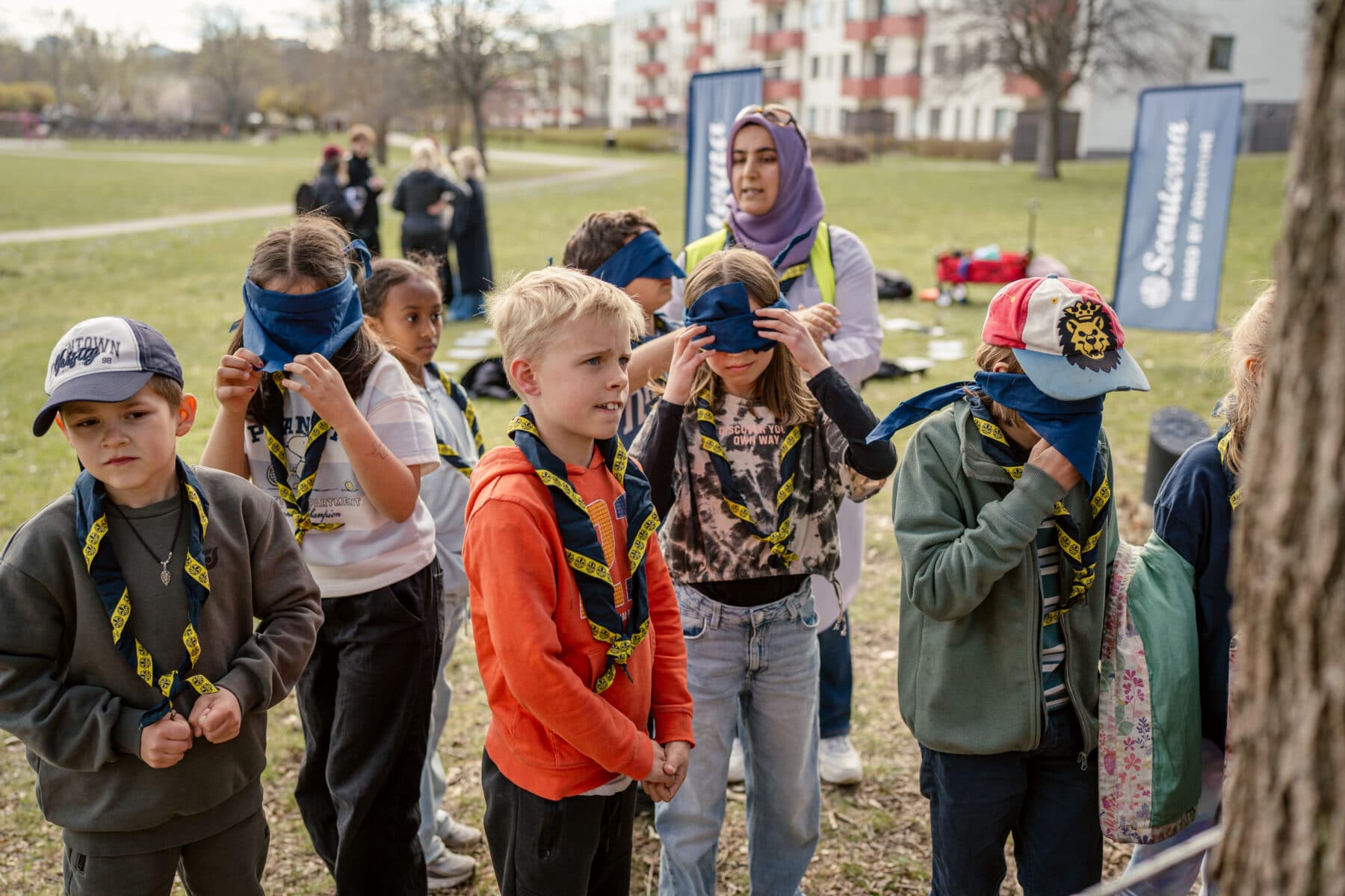 En grupp barn står utomhus på en gräsmatta, flera av dem har ögonbindlar och scoutscarfs runt halsen. I bakgrunden syns vuxna som övervakar aktiviteten samt byggnader och träd.