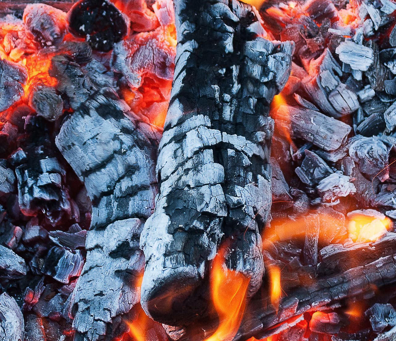 Glödande träkol med orange eldflammor och aska runt omkring.