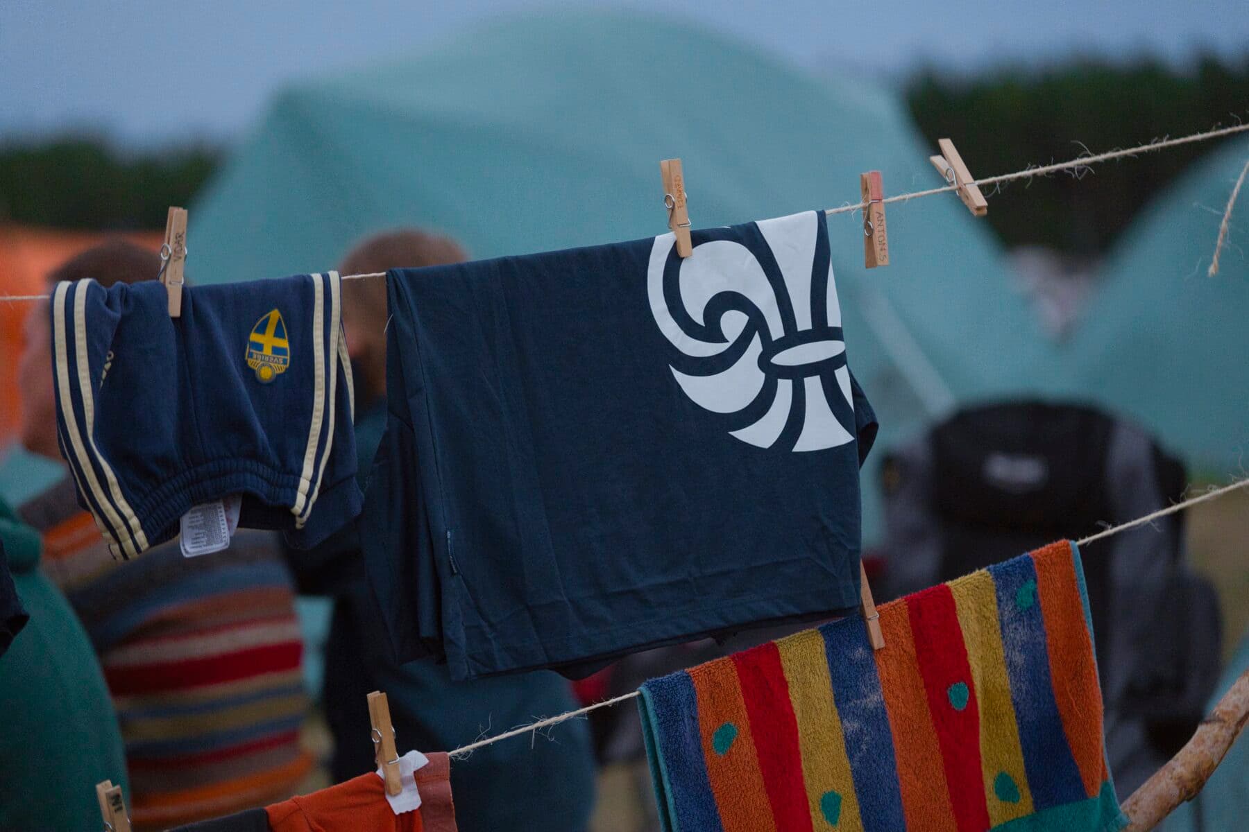 Kläder hänger på en tvättlina med klädnypor, inklusive ett par blå shorts med svensk flagga, en blå tröja med vit symbol och en färgglad handduk. Bakgrunden visar tält och suddiga personer.