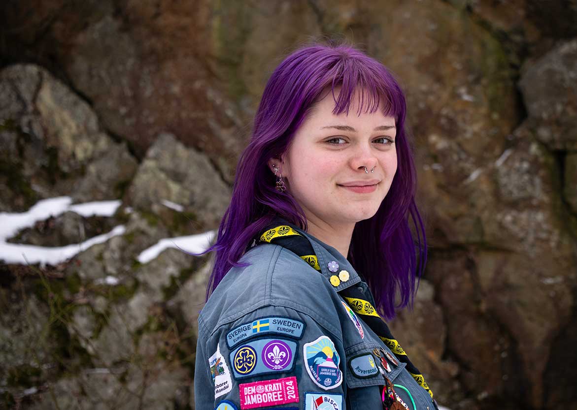 En ung person med lila hår ler mot kameran. Hen bär en scoutskjorta med flera märken, inklusive "Sverige Europa", "Demokrati Jamboree 2024" och andra scoutmärken. Bakgrunden visar en stenig yta med snöfläckar.