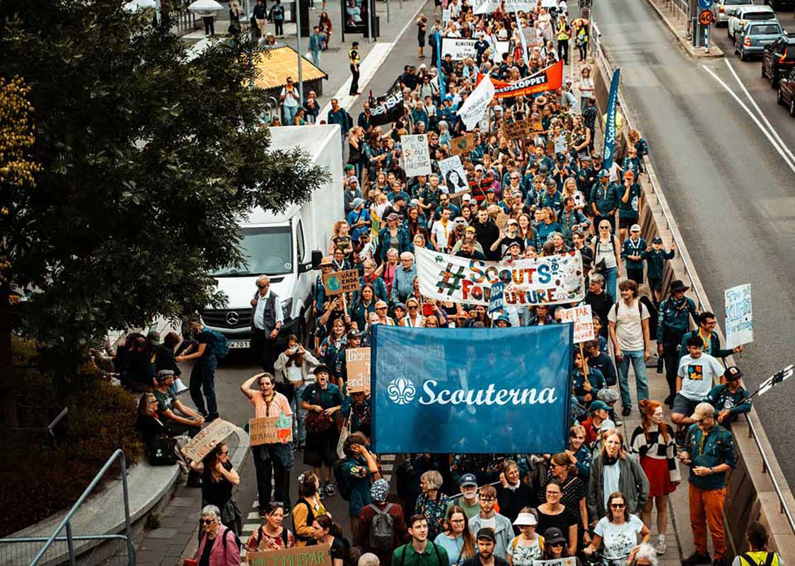 En stor grupp människor marscherar på en gata i en demonstration, med skyltar och banderoller. En blå banderoll med texten "Scouterna" samt en annan med texten "#ScoutsForFuture" syns tydligt bland deltagarna.