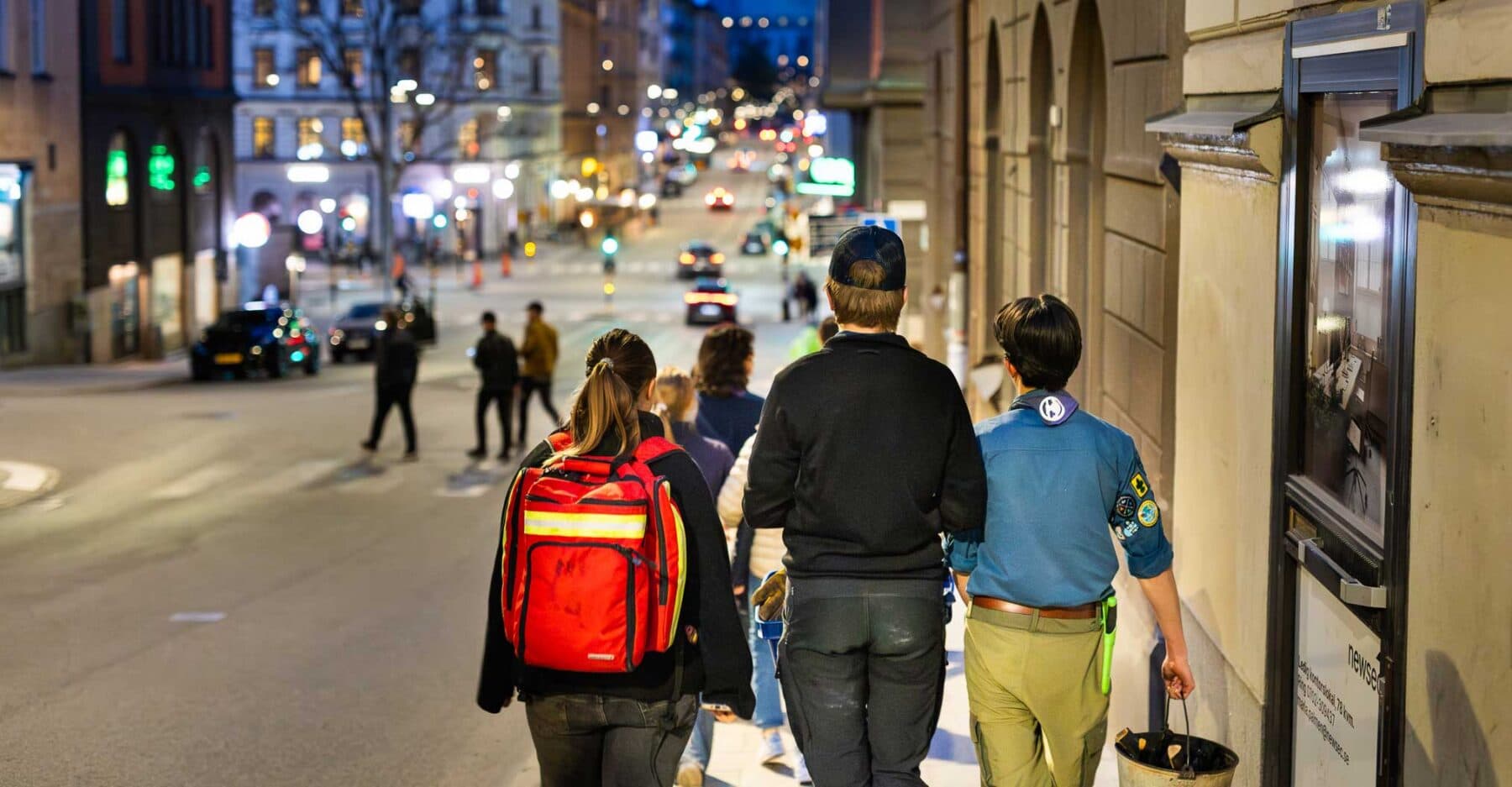 En grupp människor går längs en stadsgata på kvällen, där byggnader är upplysta och trafikljus samt bilar syns i bakgrunden. En person har en röd ryggsäck, och en annan, i scoutkläder, bär en hink.