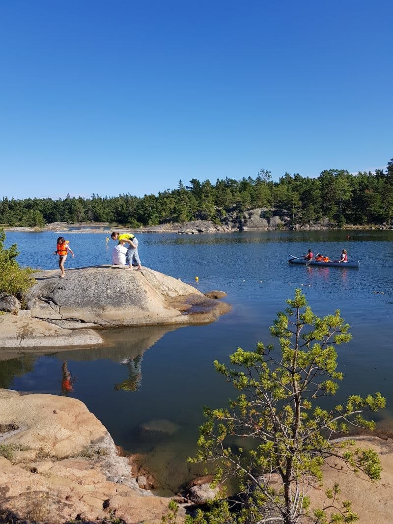 Familfer från projektet Äg din natur besöker Vässarö under en dagsutflykt. en familj står på en klippa. Några ungdomar paddlar kanot.