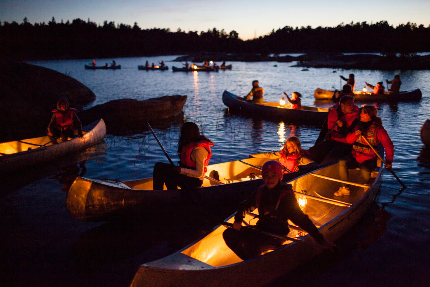 En grupp kanoter som åker genom skärgårdsmiljö i mörker. I kanoterna finns lyktor som lyser upp