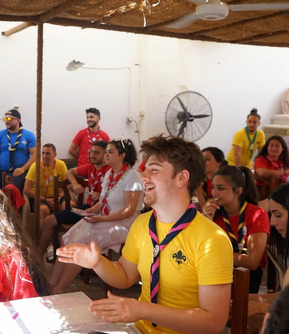 En grupp människor sitter i ett rum med vit vägg, takfläkt och halmtak. En person i gul tröja med scoutemblem gestikulerar medan andra lyssnar eller pratar.