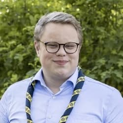 En person i ljusblå skjorta och scoutslips med gul och blå mönster står framför en grönskande bakgrund av träd och buskar.