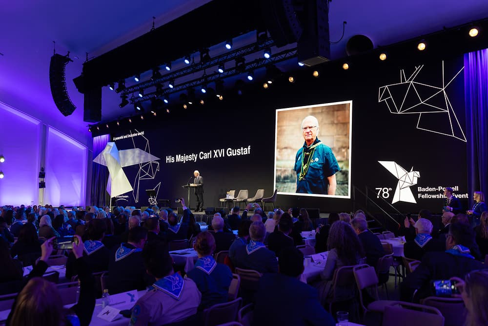 Ett stort evenemang med publik sittande vid bord. På scenen står en person och talar framför en stor skärm som visar texten "His Majesty Carl XVI Gustaf" och en bild av en person i scoutkläder. Texten "78th Baden-Powell Fellowship Event" syns också på skärmen.