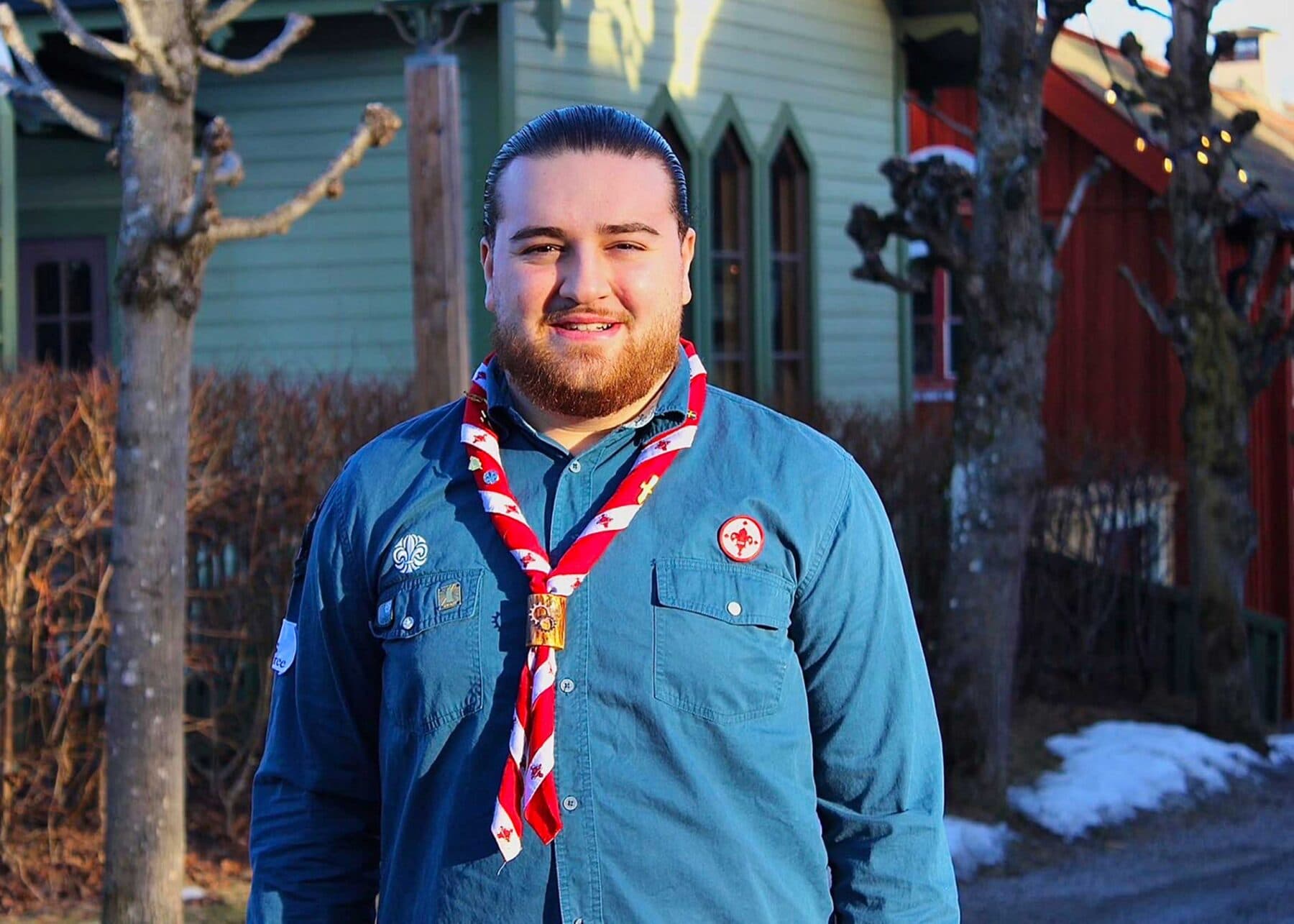 En person i en blå scoutskjorta med scoutmärken och en röd-vit halsduk står utomhus framför hus och träd i ett vinterlandskap.