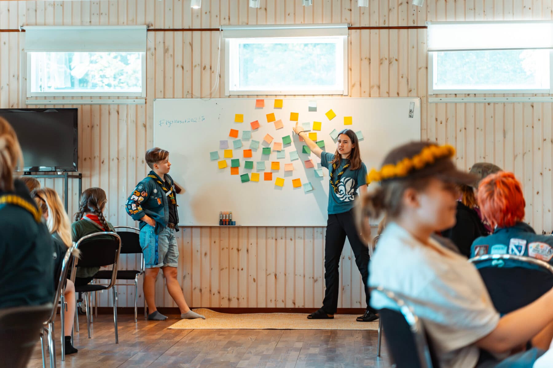 En grupp människor sitter i ett rum med träpaneler, medan en person pekar på en whiteboard fylld med färgglada lappar. Text på tavlan inkluderar "#Fyssnaset2023" och några suddiga ord.