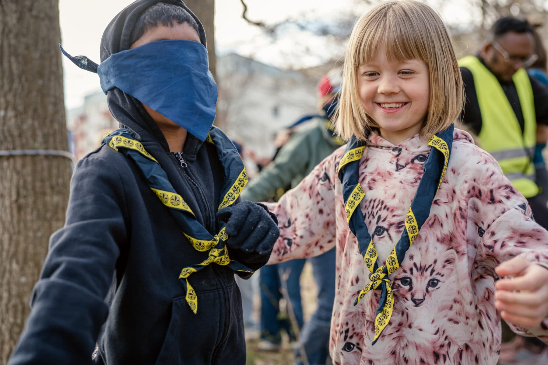 Två barn håller varandra i händerna, båda bär scoutscarfs med blå och gula detaljer. Ett barn har en tröja med kattmotiv, och bakgrunden visar personer och träd.
