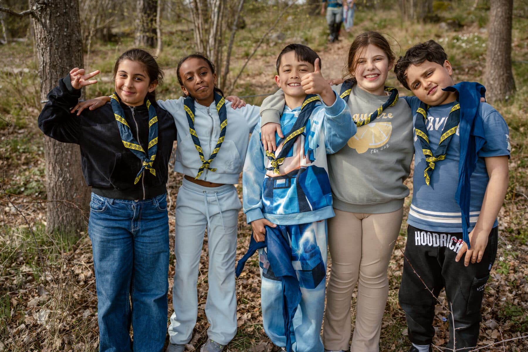 En grupp på fem barn står tillsammans i en skogsmiljö, alla med gula och blå scoutscarfar runt halsen. Barnen är klädda i olika avslappnade kläder och håller om varandra, några gör fredstecken med händerna.