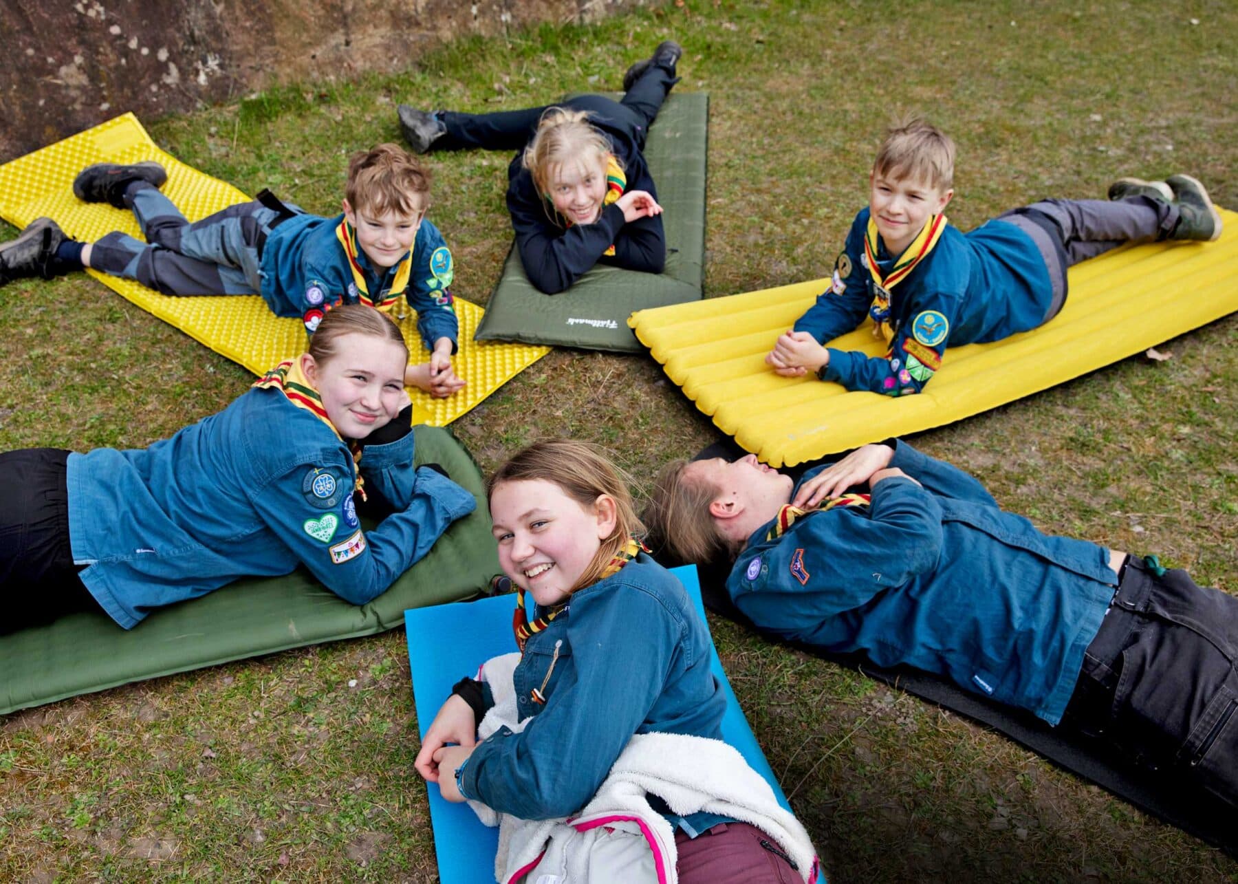 Flera personer i scoutskjortor ligger på färgglada liggunderlag på gräs, klädda i scoutuniformer med halsdukar och märken.
