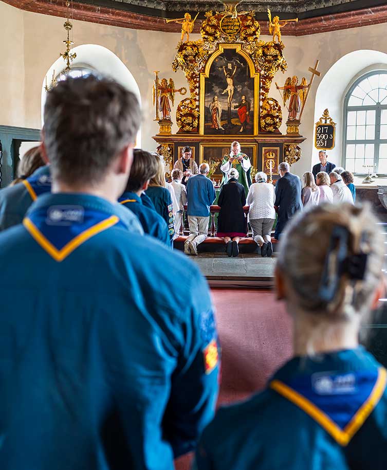 Personer i scoutuniformer står längst bak i en kyrka och tittar framåt mot en präst som leder en ceremoni vid ett dekorerat altare, medan några människor knäböjer framför altaret.