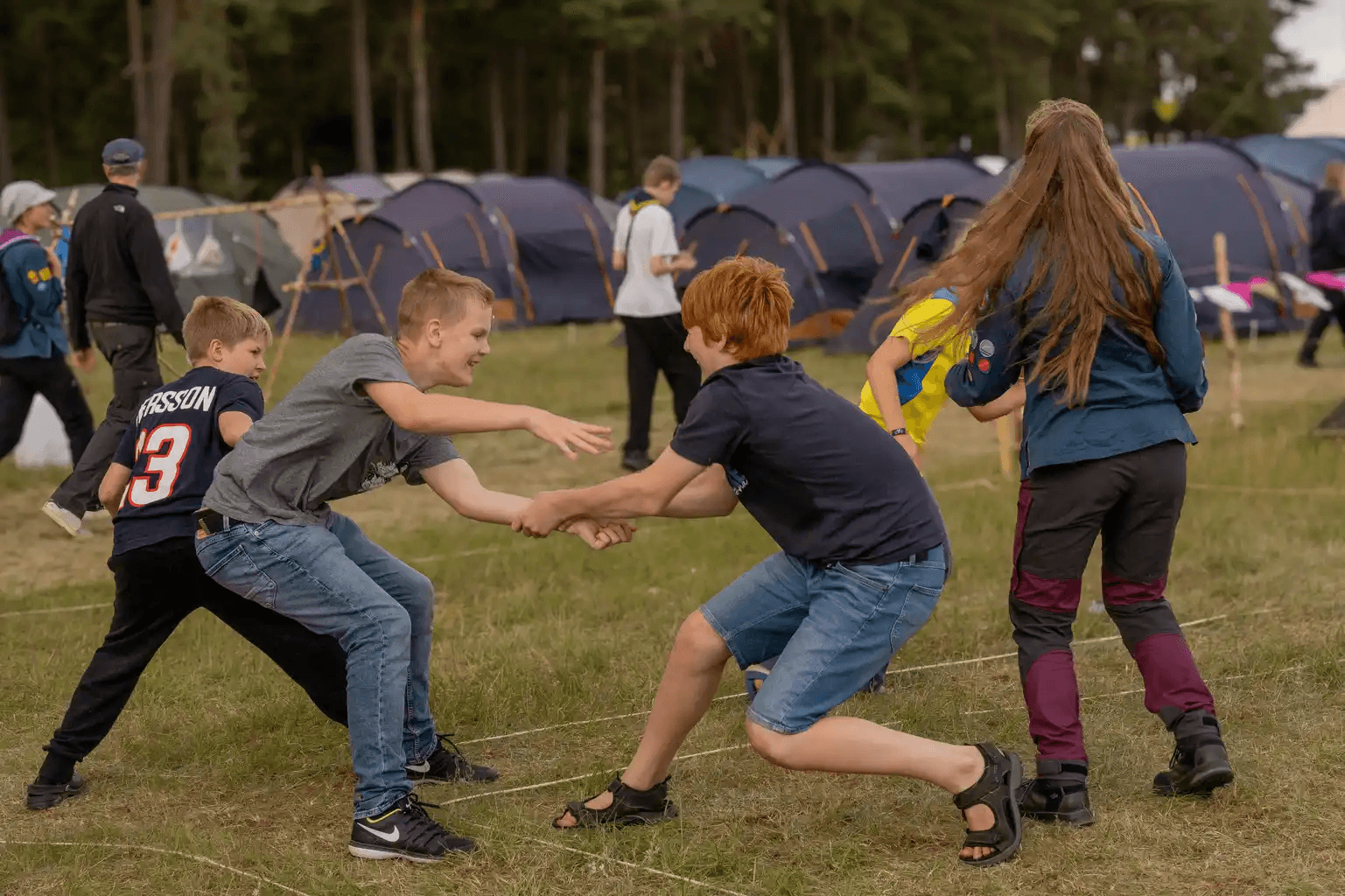 Flera barn och ungdomar leker på en gräsmatta framför tält, några håller varandras händer och verkar delta i en lek eller tävling.