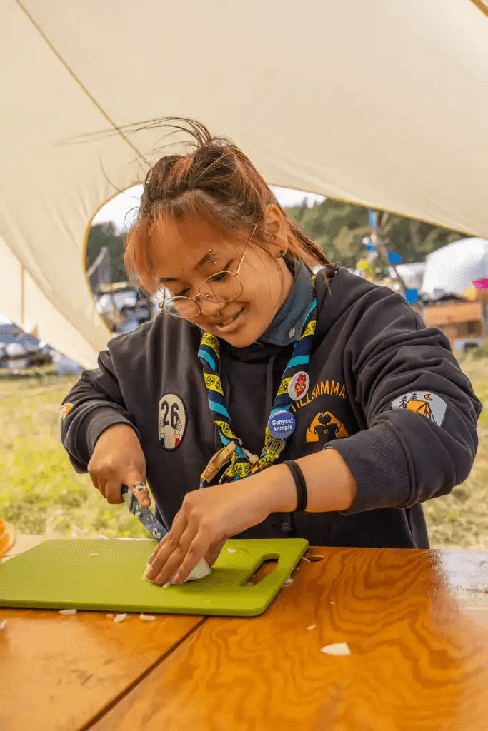 En person i scoutkläder skär en lök på en grön skärbräda vid ett träbord under ett tält.