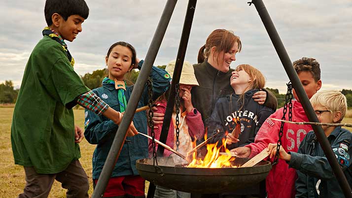 En grupp barn och en vuxen står runt en eld i en hängande eldkorg utomhus. Barnen håller träpinnar över elden och verkar glada och engagerade.