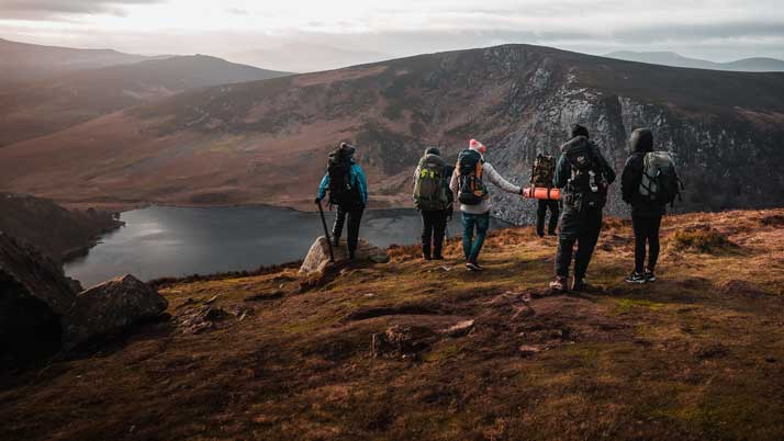 En grupp människor med ryggsäckar vandrar på en kulle med utsikt över en sjö och berg i bakgrunden under en mulen himmel.