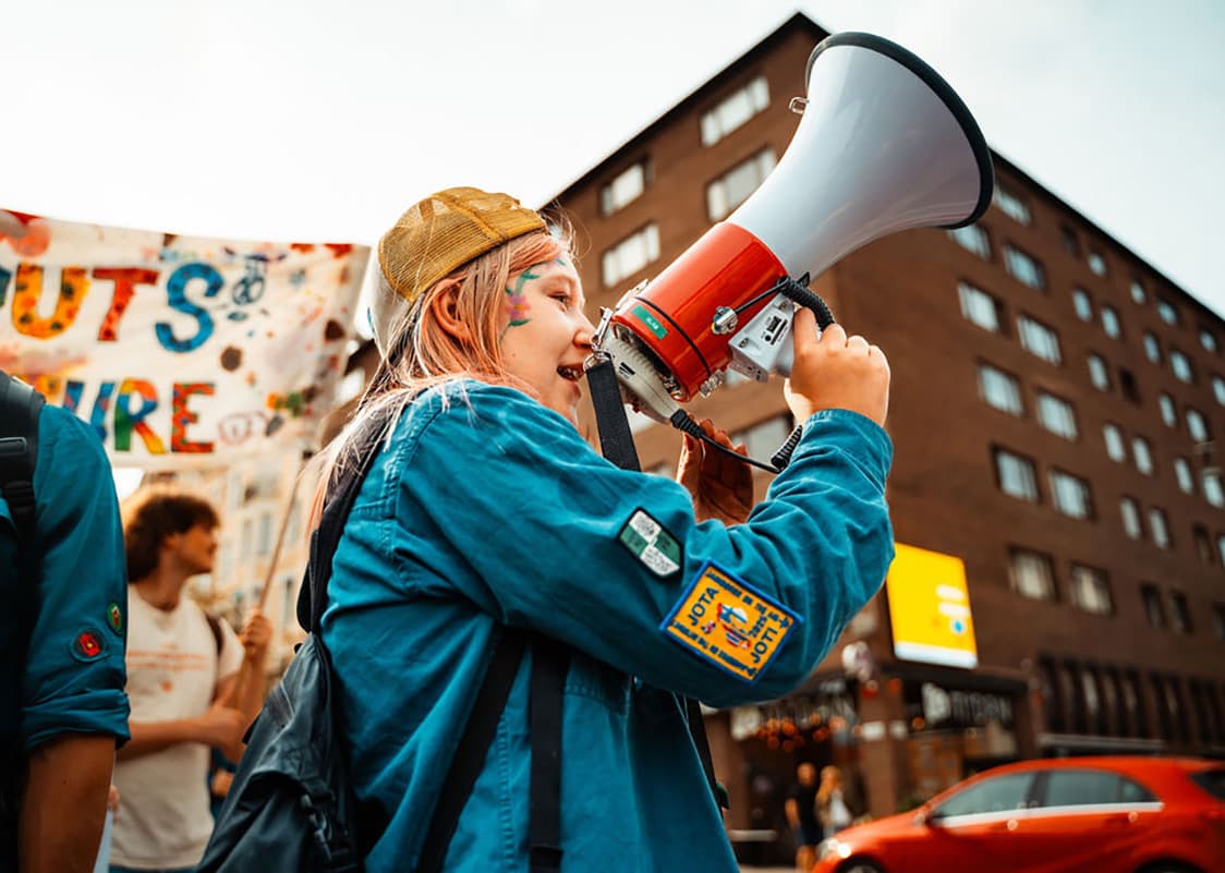 En person med blå jacka och gul keps talar i en megafon under en demonstration, med en färgglad banderoll och byggnader i bakgrunden.