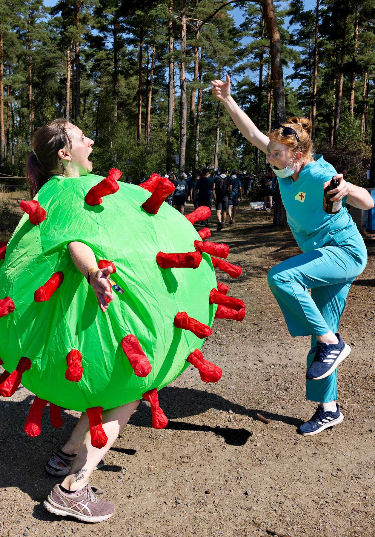 En person klädd i en grön kostym som liknar ett virus med röda utskott möter en annan person i sjuksköterskekläder som gör en dramatisk rörelse med en flaska i handen, omgiven av träd i en skogsmiljö.