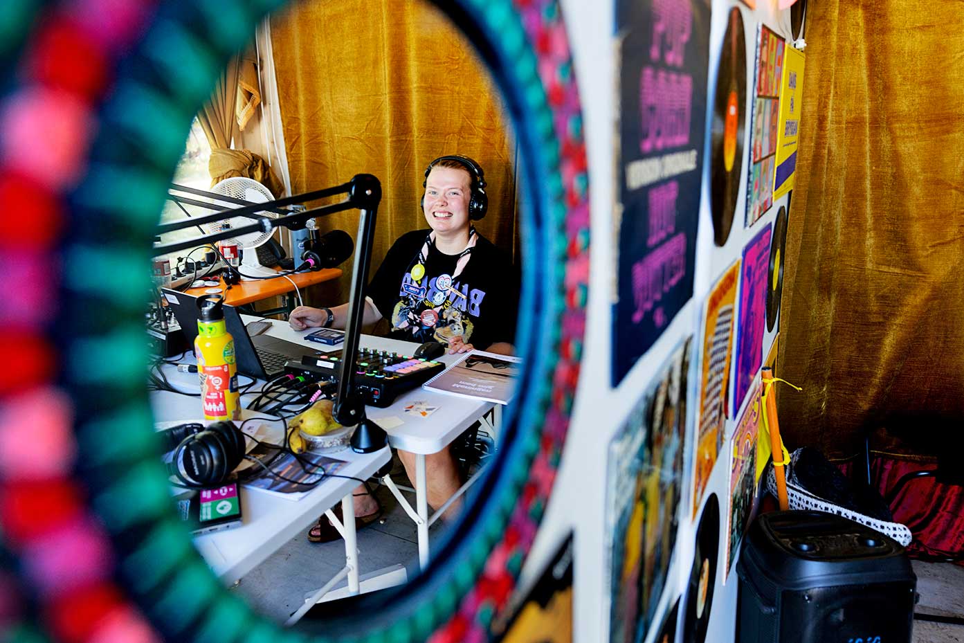 En person sitter vid ett bord i en radiostudio, omgiven av mikrofoner, hörlurar, en mixer och en laptop. Väggen bredvid har affischer och vinylskivor, och bilden är delvis inramad av ett färgglatt mönster.