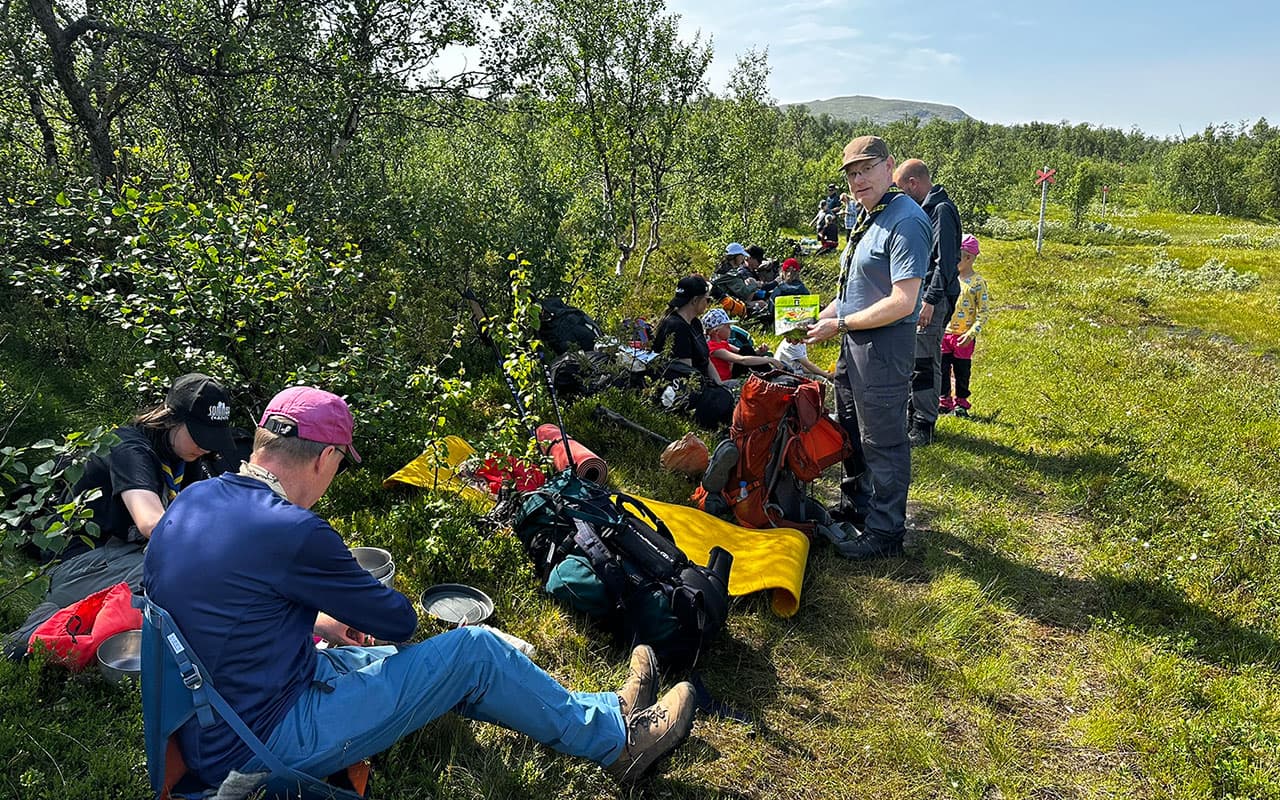 En grupp människor sitter och står på en grön äng omgiven av träd och fjäll. De har ryggsäckar, utrustning och mat med sig, och en person håller upp en förpackning.