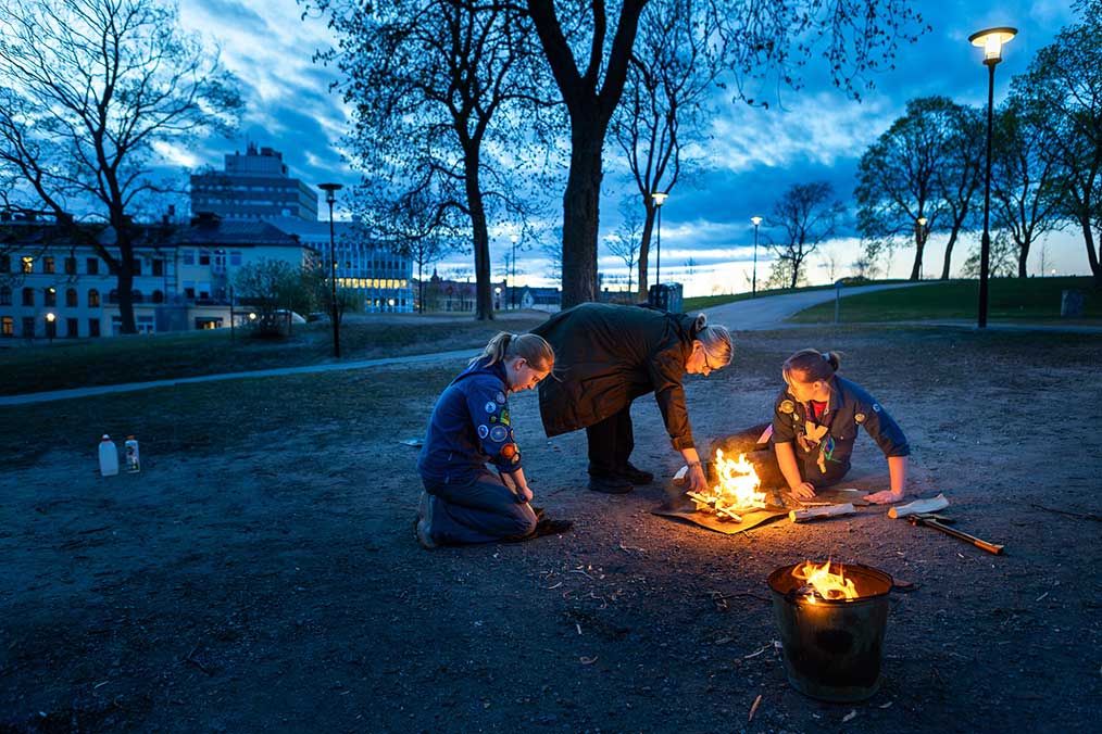 Tre personer i scoutkläder tänder en liten brasa på marken i en park under skymningen. En hink med eld står i förgrunden, och stadsmiljö med träd och gatlampor syns i bakgrunden.